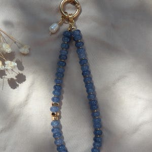 Pode incluir: Uma al&ccedil;a para telem&oacute;vel com um fio de contas azuis facetadas intercaladas com pequenos espa&ccedil;adores dourados. A al&ccedil;a prende-se a um anel e fecho de metal dourado, decorada com uma p&eacute;rola branca pendurada no ponto de fixa&ccedil;&atilde;o.
