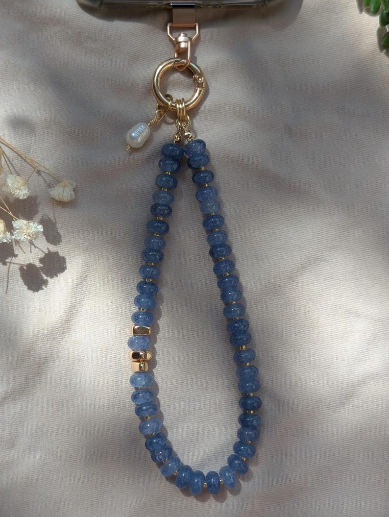 Pode incluir: Uma al&ccedil;a para telem&oacute;vel com um fio de contas azuis facetadas intercaladas com pequenos espa&ccedil;adores dourados. A al&ccedil;a prende-se a um anel e fecho de metal dourado, decorada com uma p&eacute;rola branca pendurada no ponto de fixa&ccedil;&atilde;o.