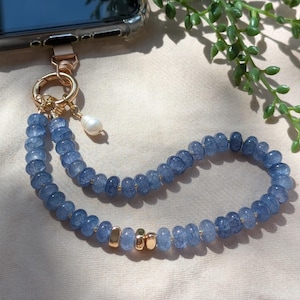 Pode incluir: Um pendente para telem&oacute;vel com um fio de contas de vidro craquelado azul, uma p&eacute;rola branca e detalhes em metal dourado. O la&ccedil;o de contas est&aacute; preso a um fecho circular dourado, ligado &agrave; extremidade superior de um smartphone.