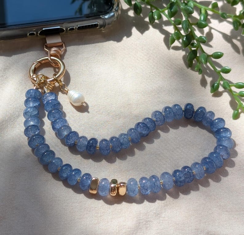 Pode incluir: Um pendente para telem&oacute;vel com um fio de contas de vidro craquelado azul, uma p&eacute;rola branca e detalhes em metal dourado. O la&ccedil;o de contas est&aacute; preso a um fecho circular dourado, ligado &agrave; extremidade superior de um smartphone.