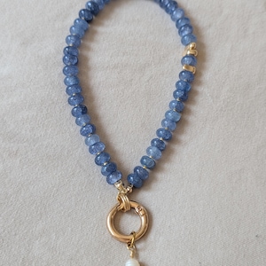 Pode incluir: Um colar de contas com pedras azuis facetadas dispostas num padr&atilde;o circular. O colar inclui detalhes em metal dourado e um fecho circular central dourado. Uma &uacute;nica conta branca semelhante a uma p&eacute;rola pende do fecho inferior.