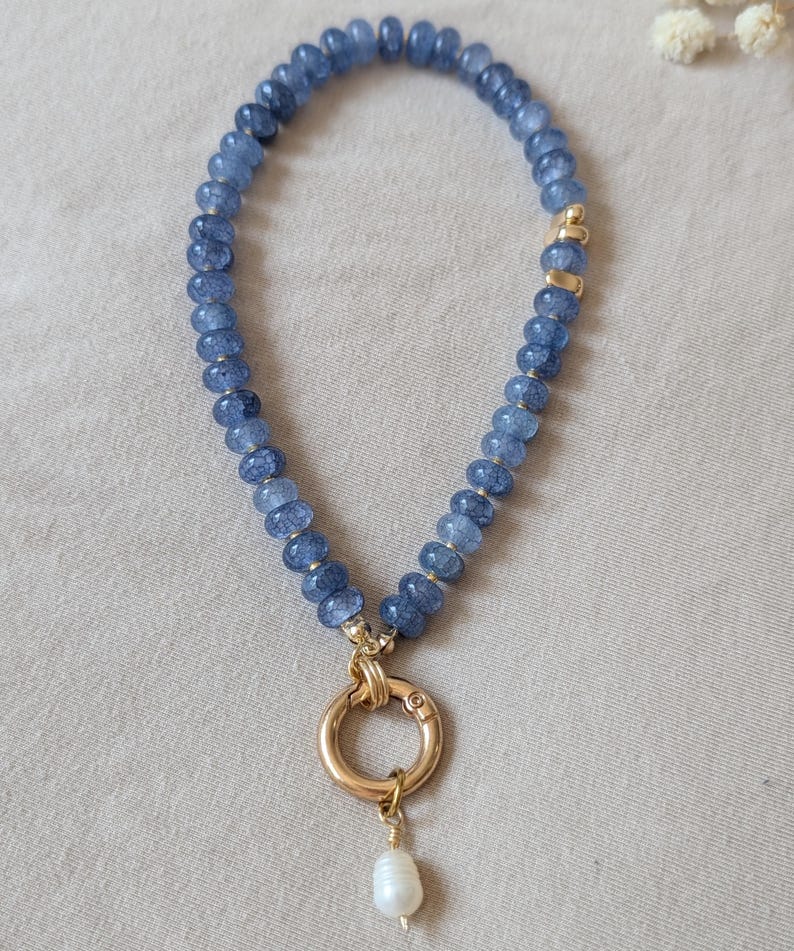 Pode incluir: Um colar de contas com pedras azuis facetadas dispostas num padr&atilde;o circular. O colar inclui detalhes em metal dourado e um fecho circular central dourado. Uma &uacute;nica conta branca semelhante a uma p&eacute;rola pende do fecho inferior.