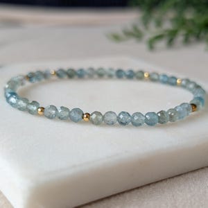 Peut inclure: Un bracelet délicat composé de perles d'aigue-marine bleu clair facettées, entrecoupées de petites perles dorées. Le bracelet est monté sur un cordon souple et repose sur une surface blanche.