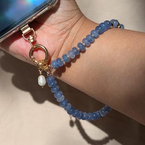 Pode incluir: Uma al&ccedil;a de pulso para telem&oacute;vel com contas azuis de textura rachada, ligadas a um fecho e anel de metal dourado. Um pequeno pendente branco semelhante a uma p&eacute;rola est&aacute; suspenso no anel dourado, dando um toque decorativo &agrave; al&ccedil;a.
