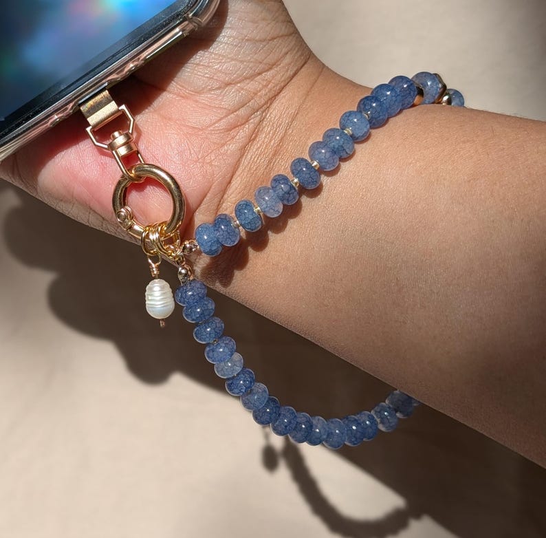 Pode incluir: Uma al&ccedil;a de pulso para telem&oacute;vel com contas azuis de textura rachada, ligadas a um fecho e anel de metal dourado. Um pequeno pendente branco semelhante a uma p&eacute;rola est&aacute; suspenso no anel dourado, dando um toque decorativo &agrave; al&ccedil;a.