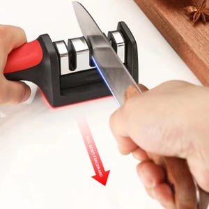 以下が含まれることがあります： 黒と赤の3スロット手動包丁研ぎ器。銀色の包丁がスロットに差し込まれており、PULL GENTLY BACKWARDというテキストが書かれた赤い矢印が、研ぐ際の動作方向を示しています。
