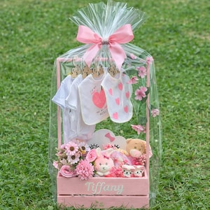 May include: A pink wooden gift basket filled with baby clothes, stuffed animals, and flowers. The basket includes white onesies with heart designs and the text "I TAKE MY HEART AND MY WORLD". The basket is wrapped in cellophane and tied with a pink bow.