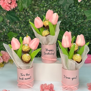 May include: Three pink floral arrangements with pink tulips and chocolate candies in pink cylindrical containers. Each container has a different message: "For the best mom!", "Happy Birthday!", and "I love you!"