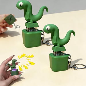 May include: Green dinosaur-shaped keychain staplers. The dinosaur has a large eye and a curved tail. The stapler is on a green square base with a silver stapler mechanism. A small square speaker is also attached to the keychain.