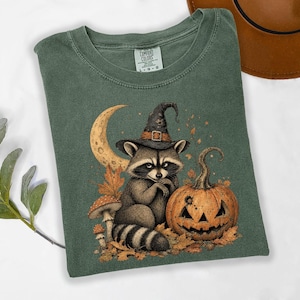 May include: A sage green t-shirt featuring a graphic print of a raccoon wearing a black witch hat, sitting next to a carved jack-o'-lantern, mushrooms, and autumn leaves under a crescent moon. The inner neck label reads Comfort Colors size L.