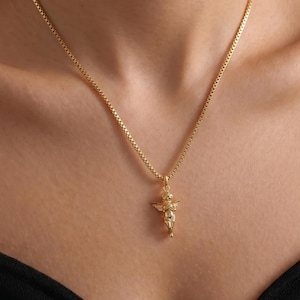 May include: A gold-colored necklace featuring a small cherub pendant. The pendant has detailed wings and is suspended from a delicate chain. The necklace is worn against a dark background.
