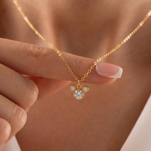 K&ouml;nnte beinhalten: Eine goldfarbene Halskette mit einem Anh&auml;nger in Form eines M&auml;usekopfes. Der Anh&auml;nger hat einen gro&szlig;en, runden, klaren Edelstein in der Mitte, mit zwei kleineren Edelsteinen dar&uuml;ber. Die Halskette ist an einer zarten Kette.
