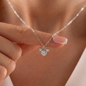 May include: A silver-toned necklace featuring a pendant shaped like a mouse head silhouette. The pendant is accented with three clear, round-cut stones, with one larger stone in the center and two smaller stones forming the ears on a delicate chain.