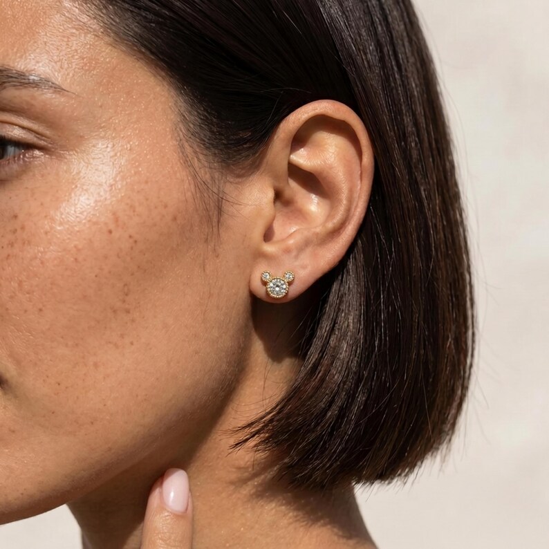 May include: A gold stud earring featuring a central round clear stone surrounded by a halo of smaller clear stones, arranged in a circular shape with two smaller circular accents at the top, resembling a stylized mouse head silhouette.
