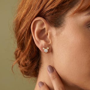 May include: A gold stud earring shaped like a mouse head, featuring a large central round clear stone surrounded by a beaded gold border, with two smaller round clear stones at the top to form ears, all set in a polished gold metal frame.