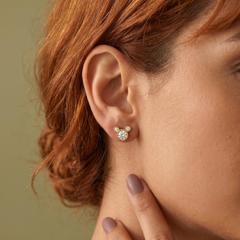 May include: A gold stud earring shaped like a mouse head, featuring a large central round clear stone surrounded by a beaded gold border, with two smaller round clear stones at the top to form ears, all set in a polished gold metal frame.