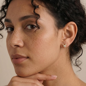 May include: A gold stud earring shaped like a mouse head silhouette, featuring a central round clear gemstone surrounded by a beaded gold border. The earring is positioned on an earlobe, showcasing a polished metallic finish and a small, circular design.