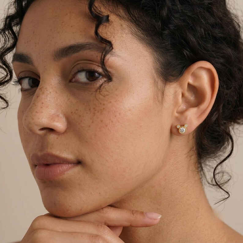 May include: A gold stud earring shaped like a mouse head silhouette, featuring a central round clear gemstone surrounded by a beaded gold border. The earring is positioned on an earlobe, showcasing a polished metallic finish and a small, circular design.