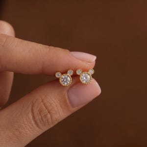 May include: A pair of gold stud earrings featuring a central round clear stone surrounded by two smaller clear stones at the top, creating a three-circle silhouette. The metal settings have a beaded texture around the edges of the stones.