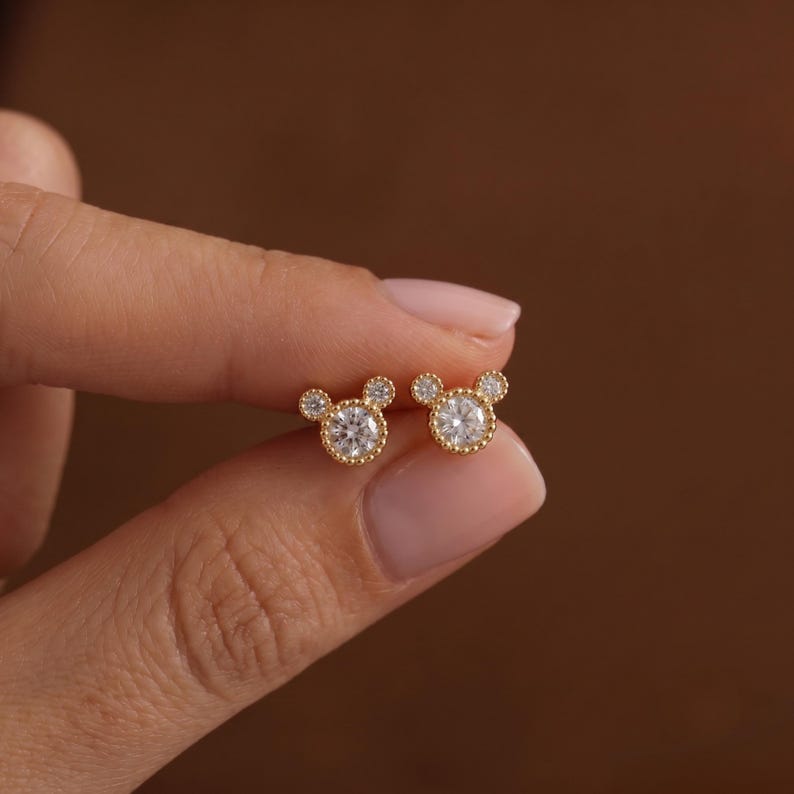 May include: A pair of gold stud earrings featuring a central round clear stone surrounded by two smaller clear stones at the top, creating a three-circle silhouette. The metal settings have a beaded texture around the edges of the stones.