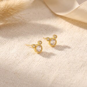 May include: A pair of gold stud earrings featuring a central round clear gemstone surrounded by a beaded metal border, with two smaller circular accents at the top, creating a silhouette resembling a mouse head shape with a post backing.