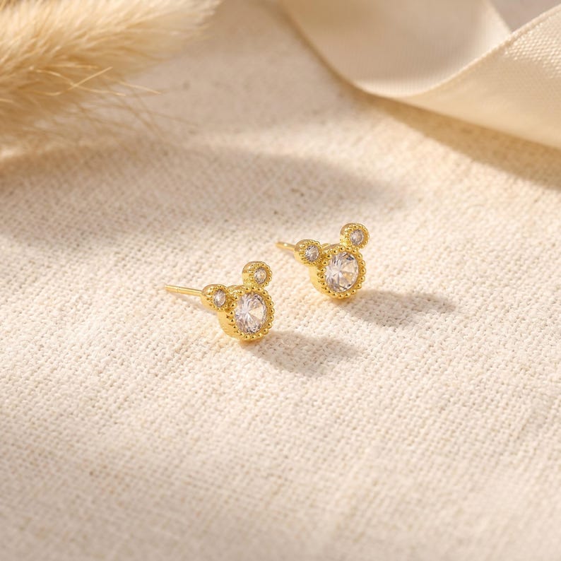 May include: A pair of gold stud earrings featuring a central round clear gemstone surrounded by a beaded metal border, with two smaller circular accents at the top, creating a silhouette resembling a mouse head shape with a post backing.