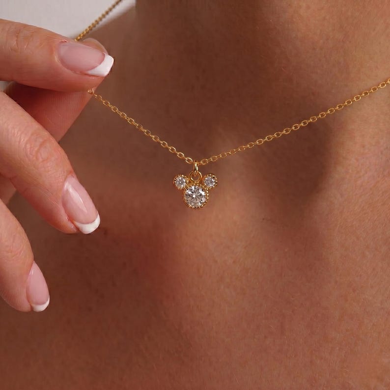 K&ouml;nnte beinhalten: Eine goldfarbene Halskette mit einem Anh&auml;nger in Form eines M&auml;usekopfes. Der Anh&auml;nger hat drei klare, runde Edelsteine. Die Halskette wird von einer Hand mit French Manicure gehalten.