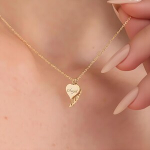 May include: A gold-colored necklace featuring a heart-shaped pendant with the word "Angel" engraved on it. The pendant also includes a wing design. The necklace is delicate and appears to be made of a fine metal.