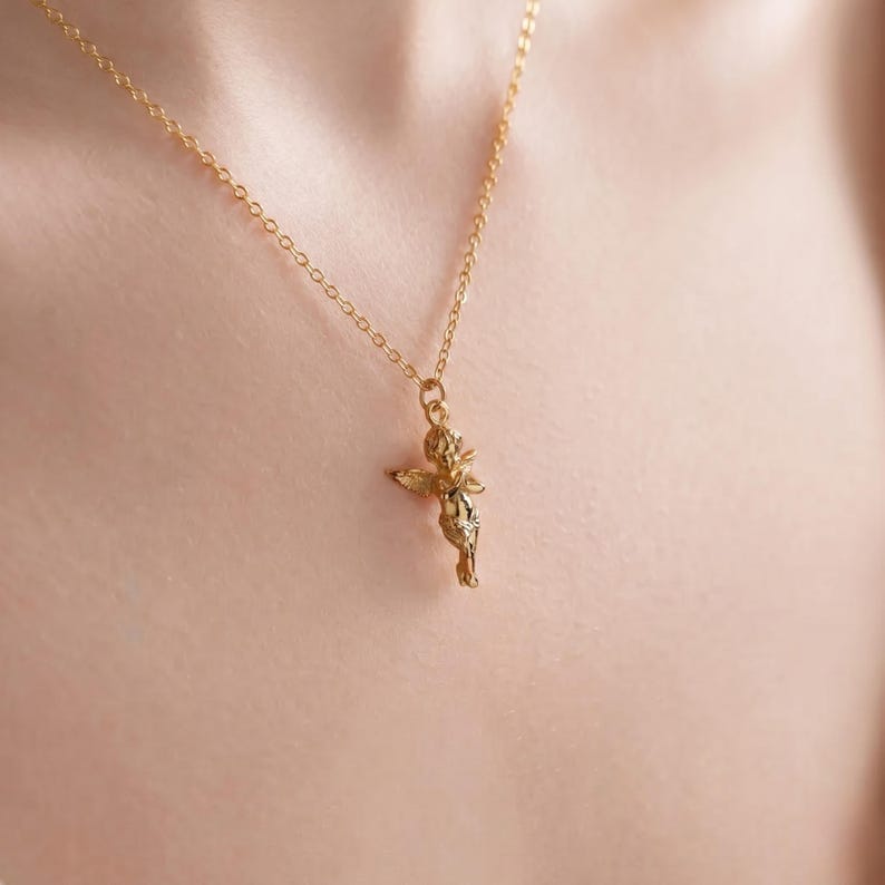 May include: A gold-colored necklace featuring a small cherub pendant. The cherub has outstretched wings and is suspended from a delicate chain. The necklace is worn against a skin-toned background, highlighting the jewelry's details.