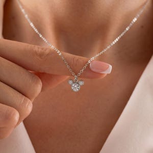 K&ouml;nnte beinhalten: Eine silberne Halskette mit einem Anh&auml;nger in Form eines M&auml;usekopfes. Der Anh&auml;nger hat einen gro&szlig;en, runden, klaren Edelstein f&uuml;r das Gesicht und zwei kleinere Edelsteine f&uuml;r die Ohren. Die Kette ist zart und silberfarben.