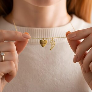 May include: A gold-colored necklace featuring a heart pendant engraved with the name "Emma" and a wing charm. The necklace is held up, showcasing the delicate chain and the two charms. The background is a soft, neutral color.