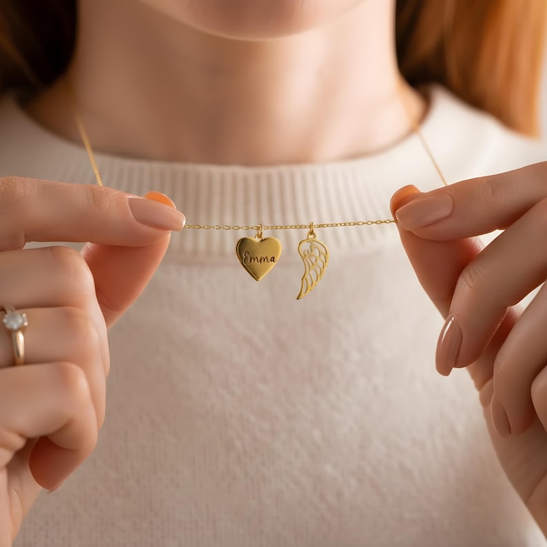 May include: A gold-colored necklace featuring a heart pendant engraved with the name "Emma" and a wing charm. The necklace is held up, showcasing the delicate chain and the two charms. The background is a soft, neutral color.
