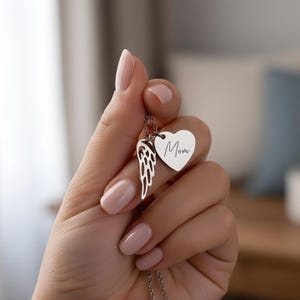 May include: A silver necklace with a heart pendant engraved with the word "Mom" and a wing charm. The necklace is held up by a hand with manicured nails. The necklace is a thoughtful gift.