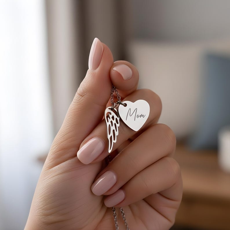 May include: A silver necklace with a heart pendant engraved with the word "Mom" and a wing charm. The necklace is held up by a hand with manicured nails. The necklace is a thoughtful gift.