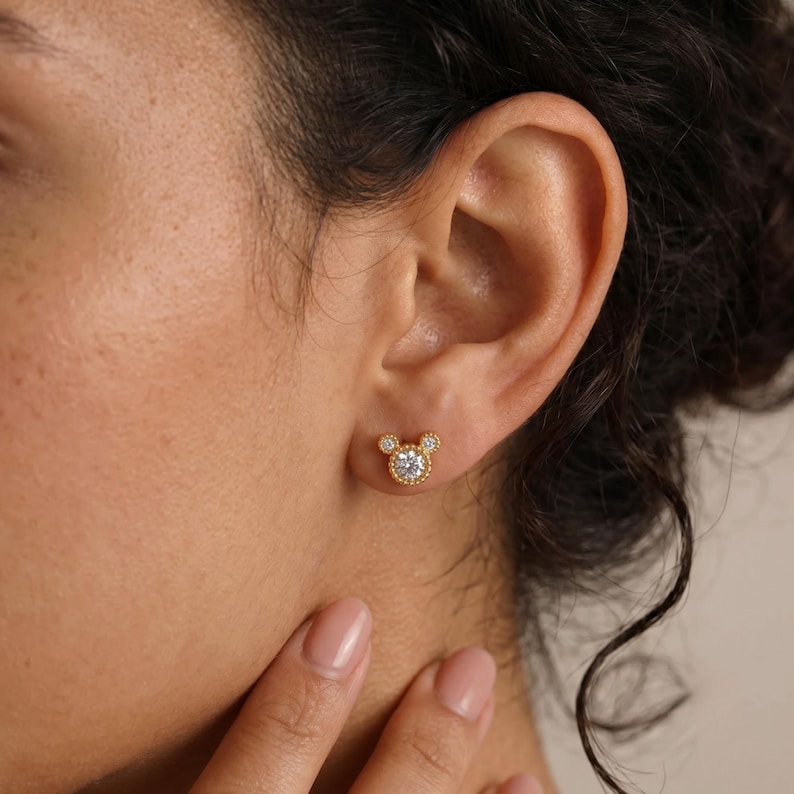 May include: A gold stud earring featuring a central round clear gemstone surrounded by two smaller clear gemstones at the top, arranged in a circular silhouette. The metal has a beaded texture along the edges, creating a distinct three-circle shape.