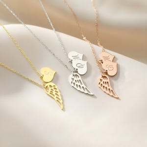 May include: Three necklaces with heart and wing pendants. One is gold, one is silver, and one is rose gold. Each necklace has a heart pendant with an initial and a wing pendant below it. The necklaces are on a light background.