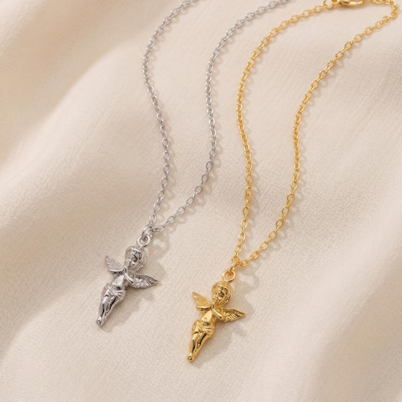 May include: Two necklaces with angel pendants. One necklace is silver-colored, and the other is gold-colored. The angel pendants have outstretched wings and are suspended from delicate chain necklaces. The background is a soft, off-white fabric.