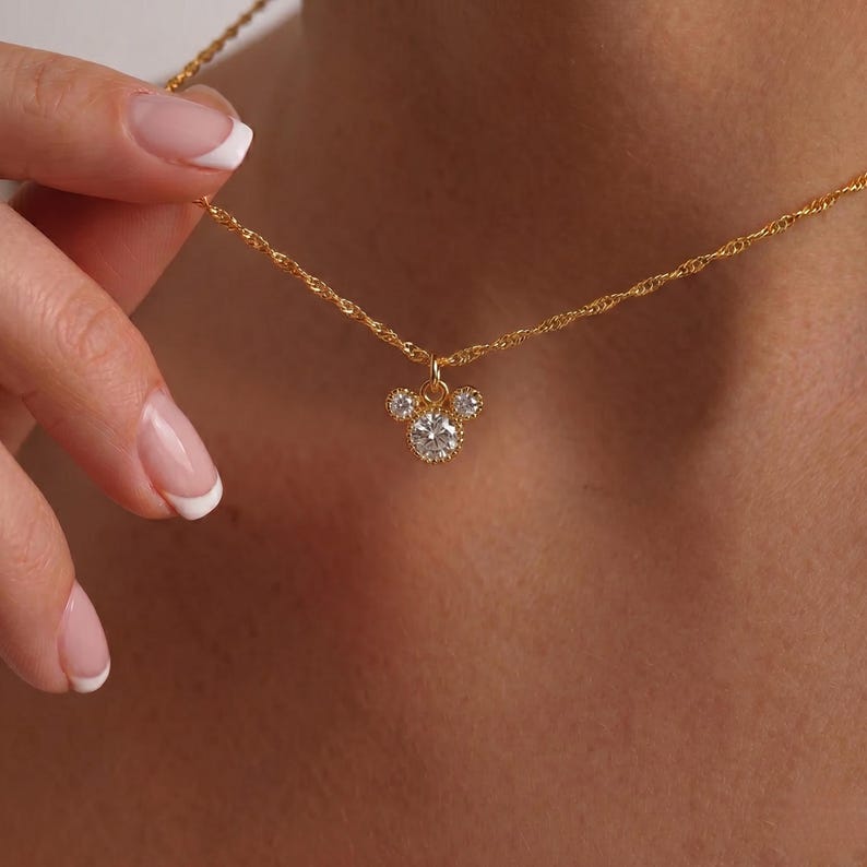 K&ouml;nnte beinhalten: Eine goldfarbene Halskette mit einem Anh&auml;nger in Form eines M&auml;usekopfes. Der Anh&auml;nger hat drei klare, runde Edelsteine. Die Halskette wird von einer Person mit gepflegten N&auml;geln gehalten.