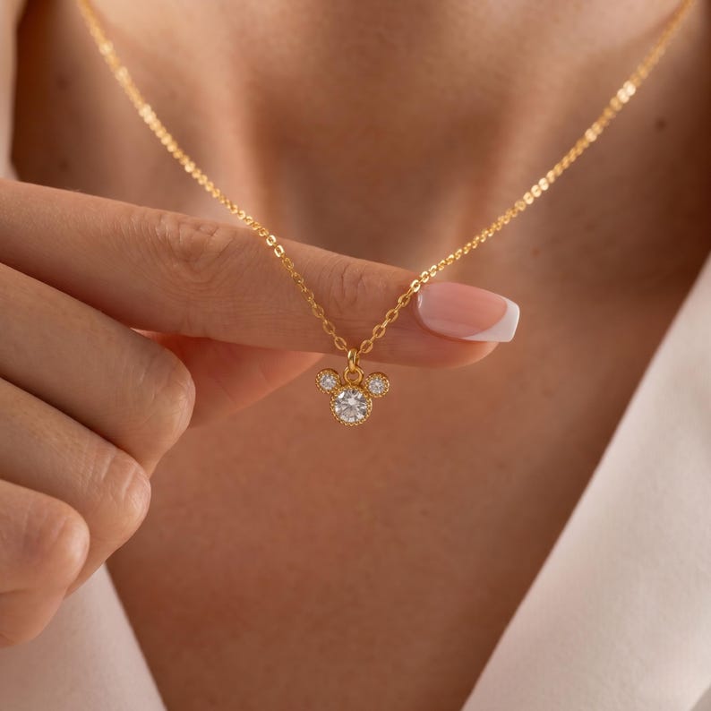 K&ouml;nnte beinhalten: Eine goldfarbene Halskette mit einem Anh&auml;nger in Form eines M&auml;usekopfes. Der Anh&auml;nger hat einen gro&szlig;en, runden, klaren Edelstein in der Mitte, mit zwei kleineren Edelsteinen dar&uuml;ber. Die Halskette ist an einer zarten Kette.