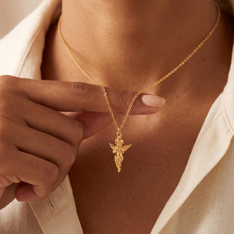 May include: A gold-colored necklace featuring a small cherub pendant. The cherub has outstretched wings and is suspended from a delicate chain. The necklace is displayed against a neutral background, highlighting the jewelry's design.