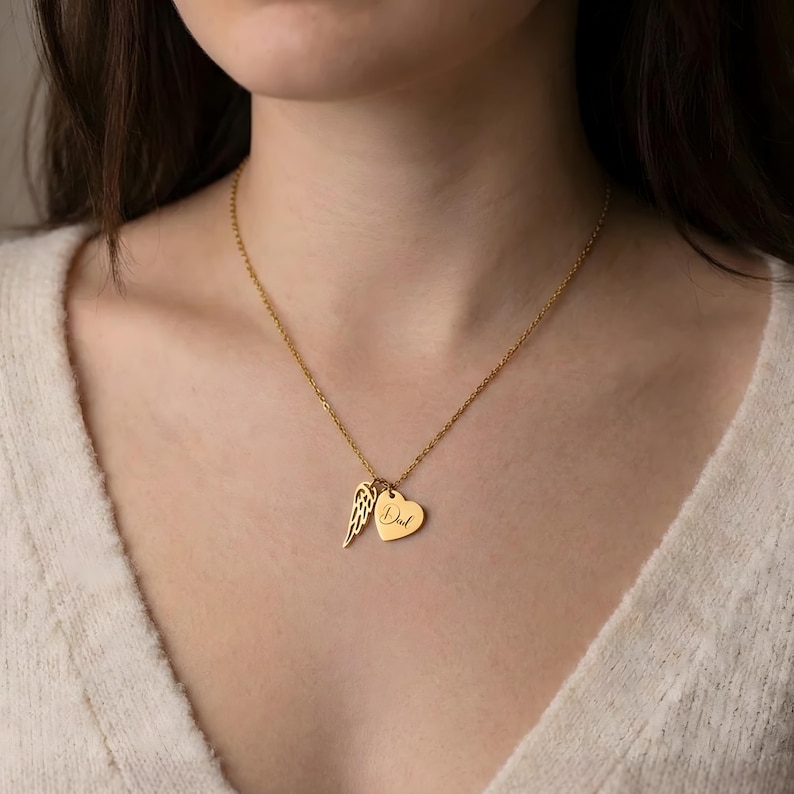 May include: Gold-colored necklace with a delicate chain. The pendant features a heart charm with the word "Dad" engraved, and a wing charm. The necklace is worn around the neck.