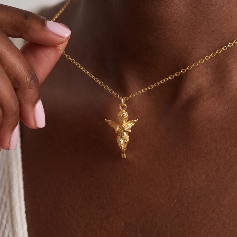May include: Gold-colored necklace featuring a small cherub pendant. The cherub has outstretched wings and clasped hands. The chain is delicate and the pendant is intricately detailed. The necklace is shown against a dark background.