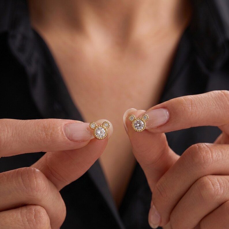 May include: A pair of gold stud earrings featuring a central round clear stone surrounded by a beaded metal border, with two smaller round clear stones positioned at the top to create a circular silhouette resembling a stylized mouse head shape.