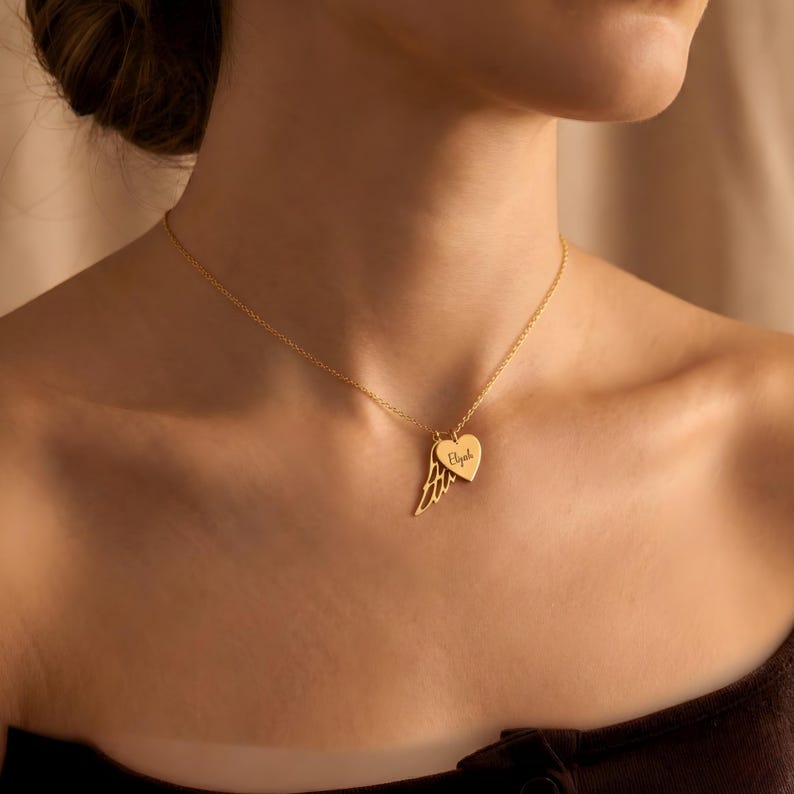 May include: A gold-colored necklace featuring a heart-shaped pendant with an engraved name and a wing charm. The delicate chain and charms are displayed against a neutral background, highlighting the jewelry's design and craftsmanship.