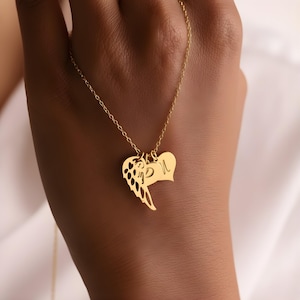 May include: A gold-colored necklace featuring three pendants: an angel wing, a heart, and a heart-shaped pendant with the letter "L". The necklace is displayed against a neutral background.
