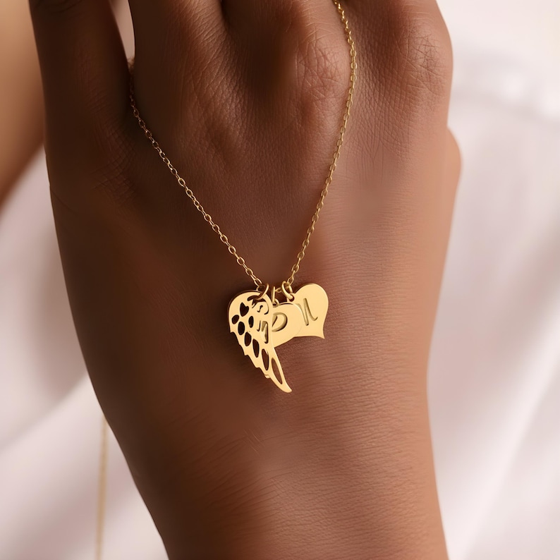 May include: A gold-colored necklace featuring three pendants: an angel wing, a heart, and a heart-shaped pendant with the letter "L". The necklace is displayed against a neutral background.