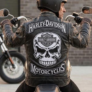 Op de afbeelding: Een persoon draagt een zwarte helm en een leren jas met Harley-Davidson-patches, waaronder een schedelafbeelding. De persoon zit op een motorfiets, met het stuur zichtbaar. De jas is donkerbruin.