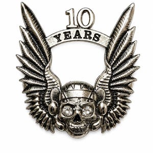 Puede incluir: Un broche de metal plateado que presenta una calavera central con casco y grandes alas que se extienden hacia arriba a ambos lados. La calavera tiene ojos de cristal transparente. Sobre ella, una banda curva muestra el texto 10 YEARS.