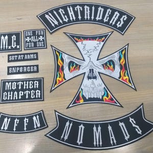 Puede incluir: Una colección de parches bordados en blanco y negro. Los parches incluyen textos como "NIGHTRIDERS", "NOMADS", "MOTHER CHAPTER" y "M.C.". Un parche grande presenta una calavera dentro de un diseño de cruz con llamas.