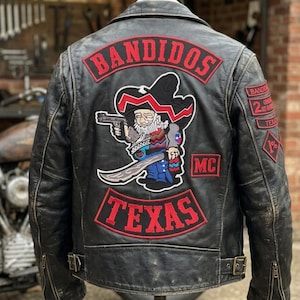 Puede incluir: Chaqueta de cuero negra de motociclista con parches. La parte trasera presenta un parche grande con una figura de dibujos animados sosteniendo una pistola y una espada, con las palabras "BANDIDOS" y "TEXAS" en rojo. Parches adicionales en las mangas.